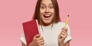 Rear view of joyful glad smiling woman with happy expression, holds red book and pencil in both hands, dressed in white t shirt, poses against pink background. People, emotions and education concept